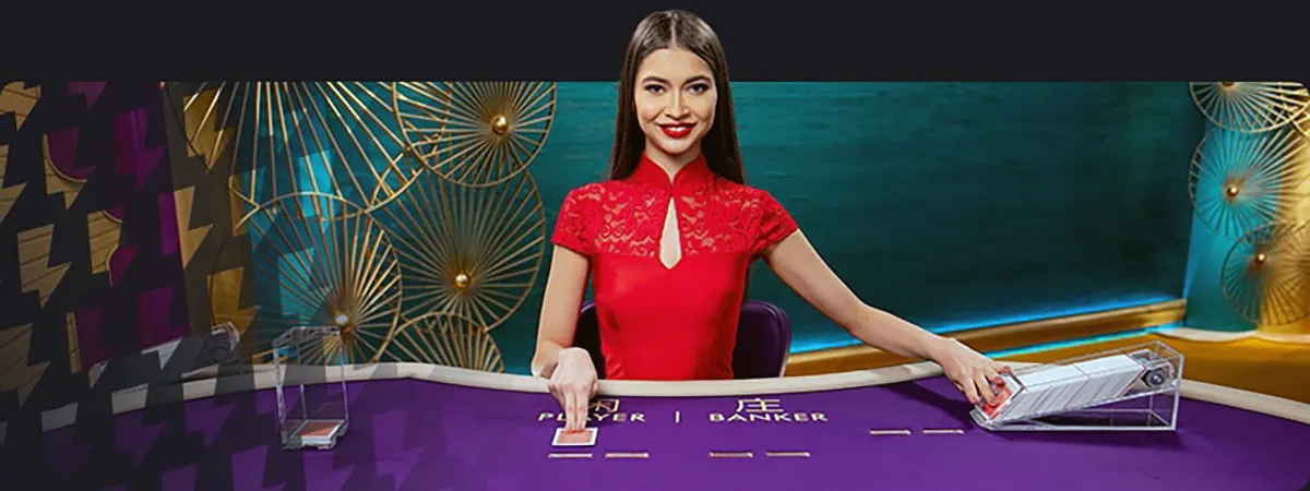 Smiling live casino dealer in a red dress sits behind a purple baccarat table labeled “Player” and “Banker,” with card shoe and discard tray on the sides and decorative gold wall accents in the background.